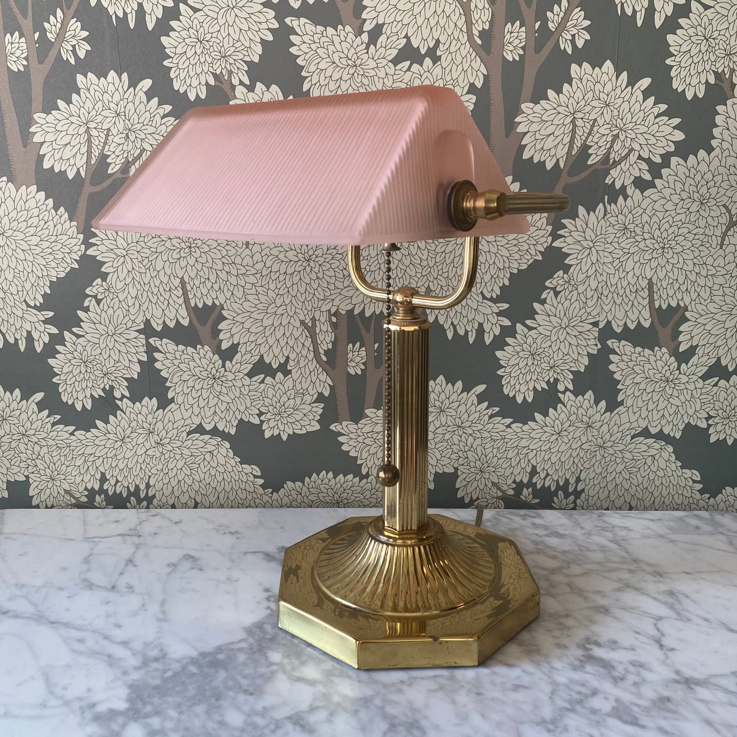 Vintage brass bankers table lamp with pink glass shade on a marble surface with floral wallpaper background.