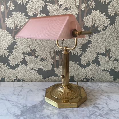 Vintage brass bankers table lamp with pink glass shade on a marble surface with floral wallpaper background.