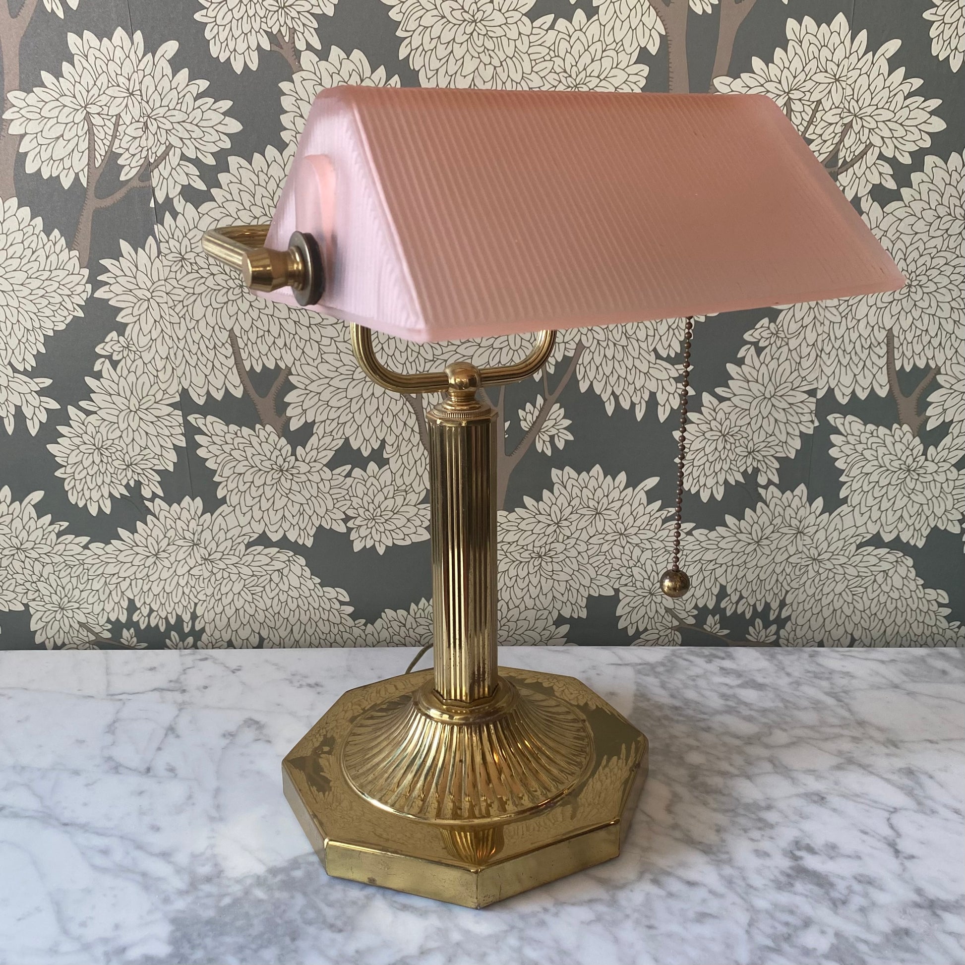 Vintage brass desk lamp with pink glass shade on a marble surface with floral wallpaper.