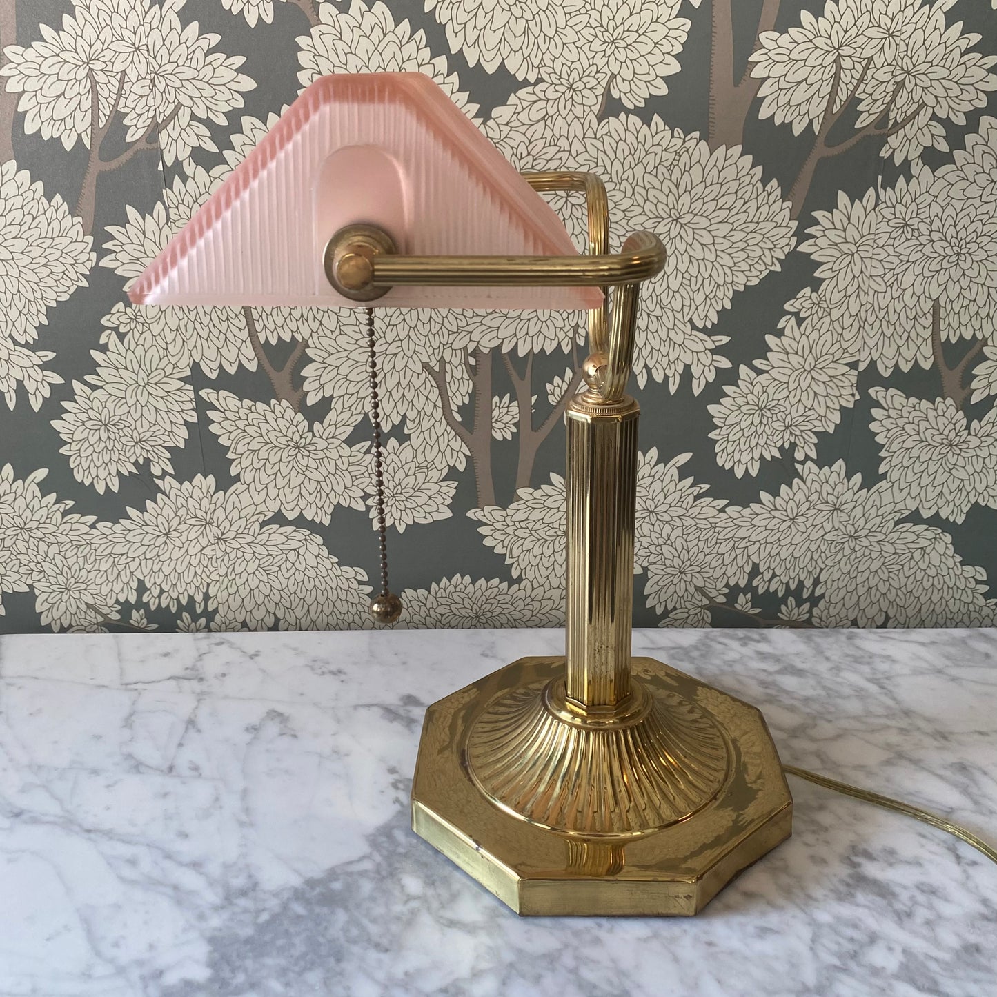 Vintage brass bankers desk lamp with a pink shade on a marble surface with floral wallpaper in the background