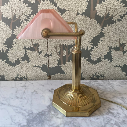 Vintage brass bankers desk lamp with a pink shade on a marble surface with floral wallpaper in the background